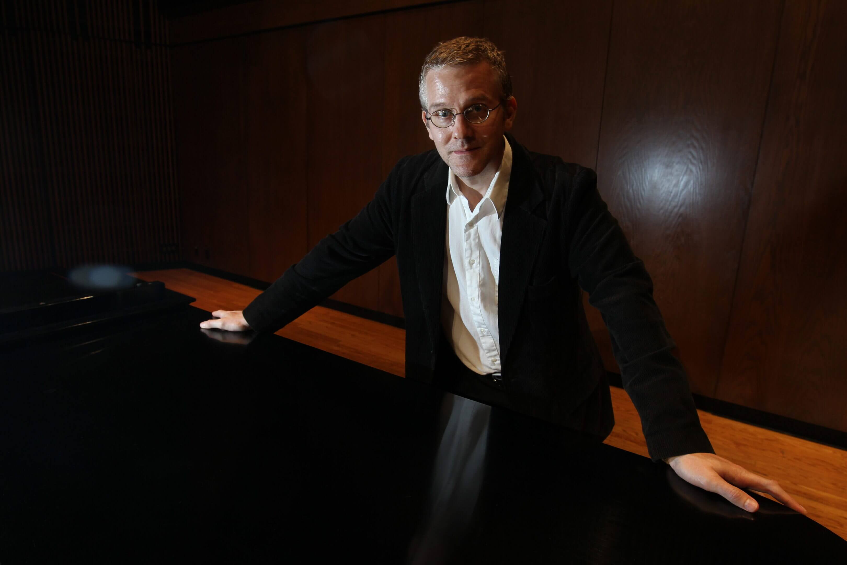 Man in a black suit playing a grand piano in a dimly lit room.